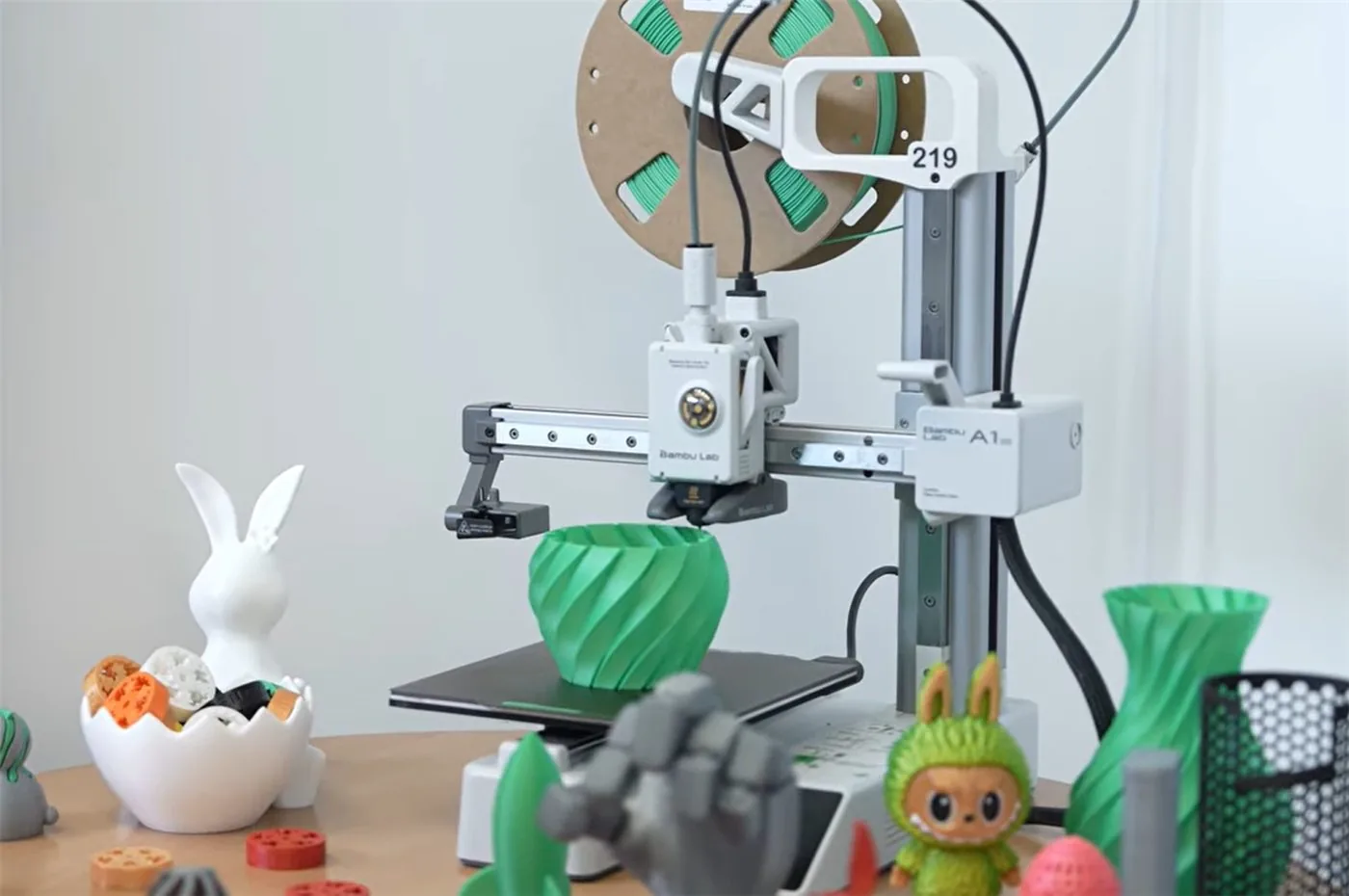 Bambu Lab A1 mini 3D nyomtató nyomtatás közben, körülötte kinyomtatott figurák, vázák és játékok