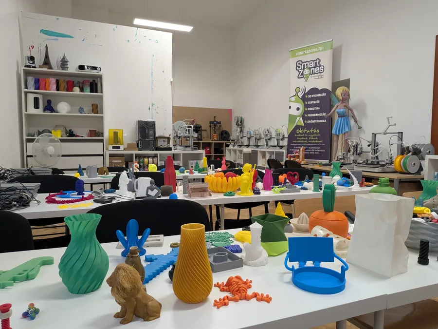 Asztalok tele 3D nyomtatott vázákkal, figurákkal és játékokkal, a háttérben 3D nyomtatók